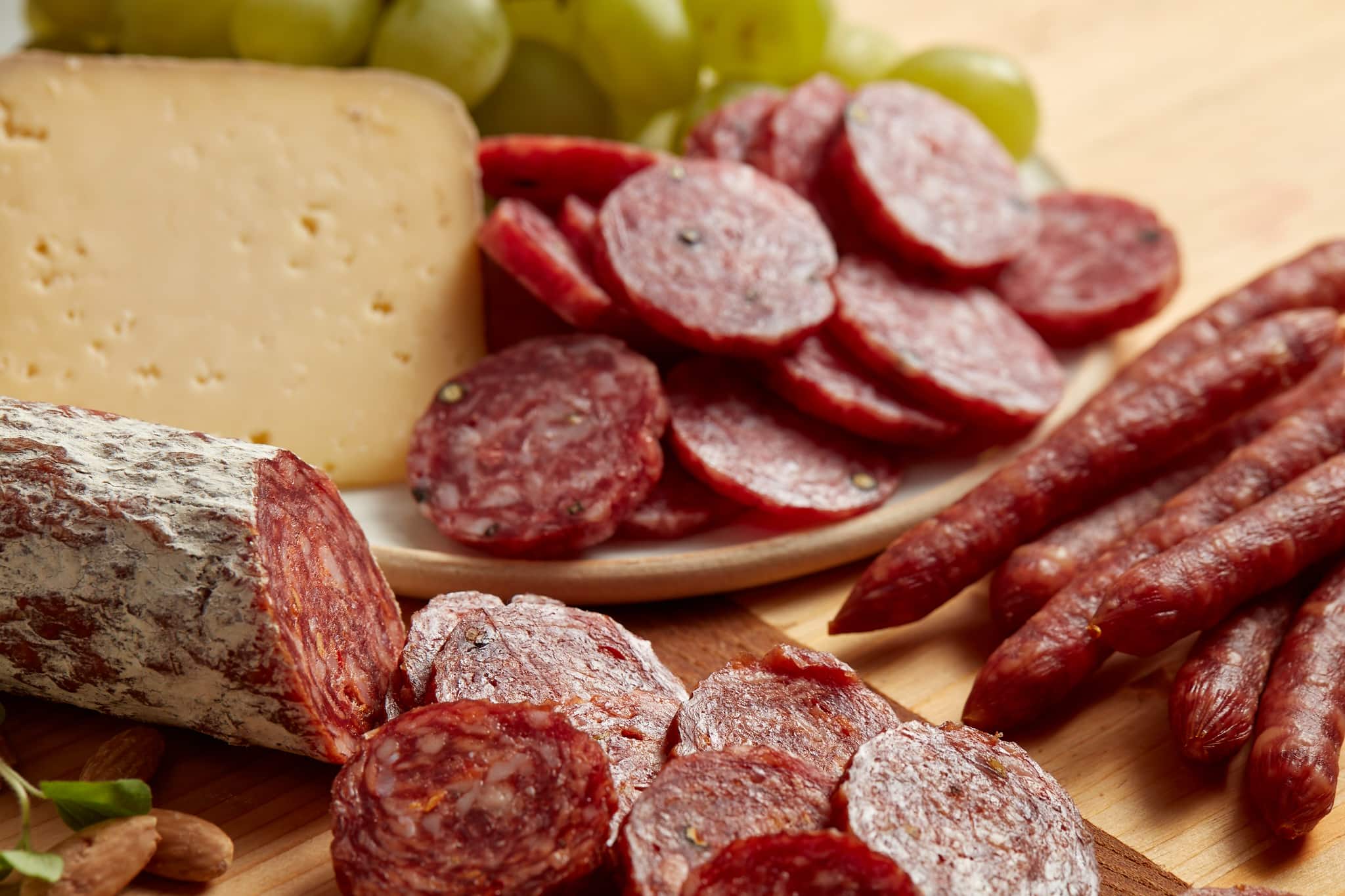 Different types of cured Olli salami on a board and plate with cheese and grapes